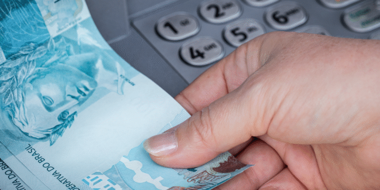 Mão retirando notas de cem reais de caixa eletrônico no Brasil.