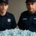 Dois policiais posando ao lado de uma grande pilha de cédulas de 100 reais, simbolizando o pagamento de bônus extra para policiais e guardas municipais.