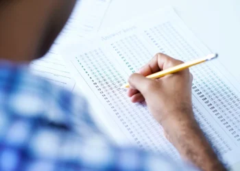 Pessoa revisando a chave de respostas de um exame, com lápis, indicando o processo de revisão para garantir acertos.