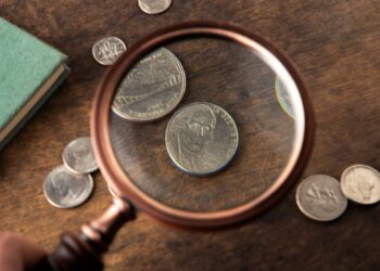 Uma lupa em close-up focada em moedas americanas, como um níquel com o perfil de Thomas Jefferson, sobre uma mesa de madeira.
