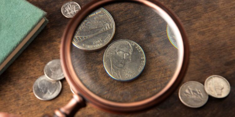 Uma lupa em close-up focada em moedas americanas, como um níquel com o perfil de Thomas Jefferson, sobre uma mesa de madeira.