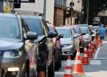 Carros parados em fila em frente a um posto de gasolina, com cones de sinalização na calçada.