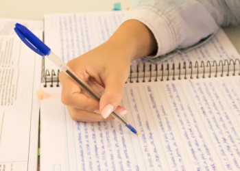 Mão de estudante escrevendo em um caderno com caneta azul, ao lado de livros e anotações, simbolizando a preparação para redação do Enem.