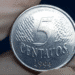 Close-up de uma moeda brasileira de 5 centavos do ano de 1994, segurada por uma mão com luva.