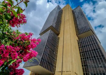 Edifício do Banco Central do Brasil em Brasília, com flores cor-de-rosa em primeiro plano, ilustrando a nova política de restrição de transferências para contas suspeitas de atividades ilícitas.
