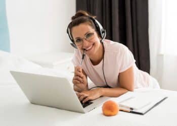 Mulher estudando de casa com notebook, fones de ouvido e anotações, representando a carreira de educador on-line.