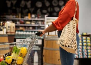 Consumidora consciente com sacola ecológica reutilizável empurrando carrinho com frutas e vegetais frescos em supermercado moderno.