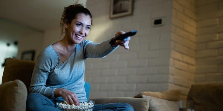 Mulher assistindo transmissão ao vivo em casa com controle remoto e pipoca aproveitando streaming .