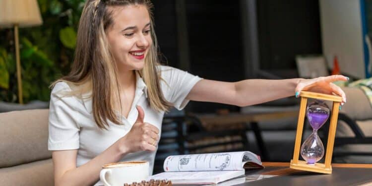 Mulher sorridente com ampulheta controlando tempo de estudo com livros e café na mesa