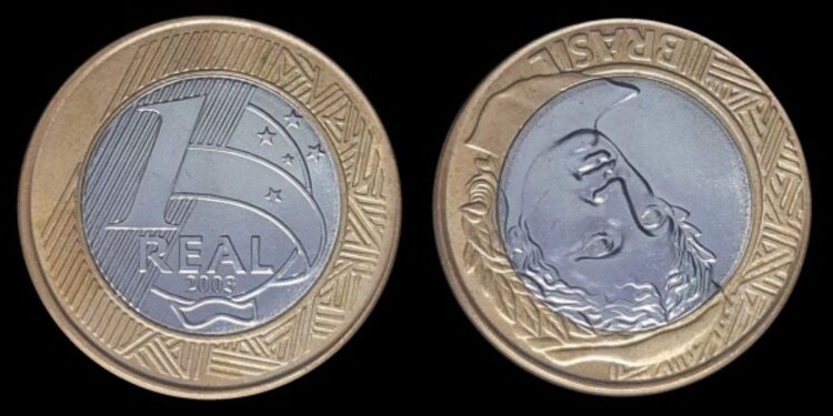 A imagem exibe a moeda de 1 real de 2003 com o erro de reverso invertido, mostrando o lado com o valor e o lado com a Efígie da República virado de cabeça para baixo.