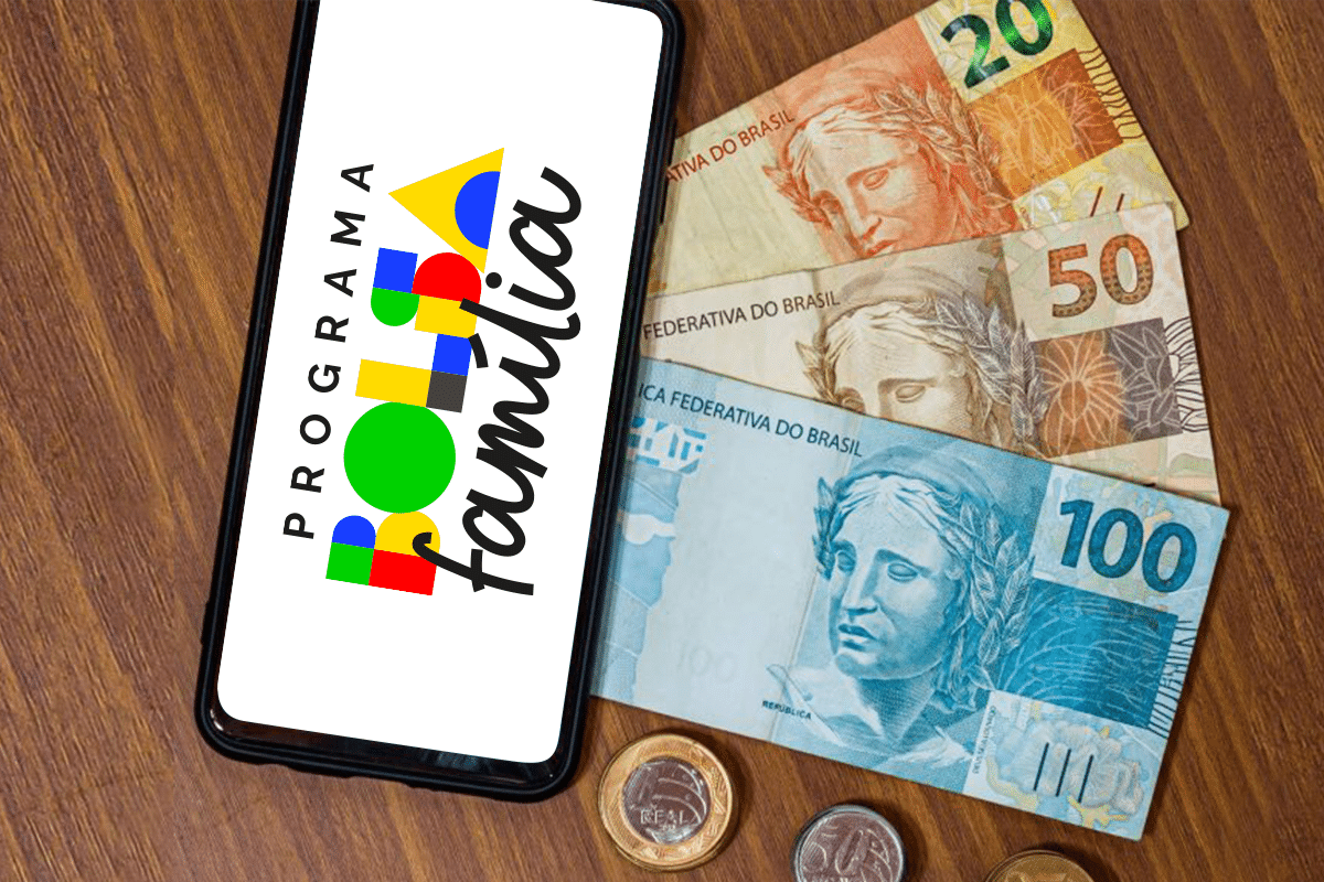 Smartphone exibindo o logo do Programa Bolsa Família, com cédulas de R, R e R0, além de moedas em cima de uma mesa de madeira.