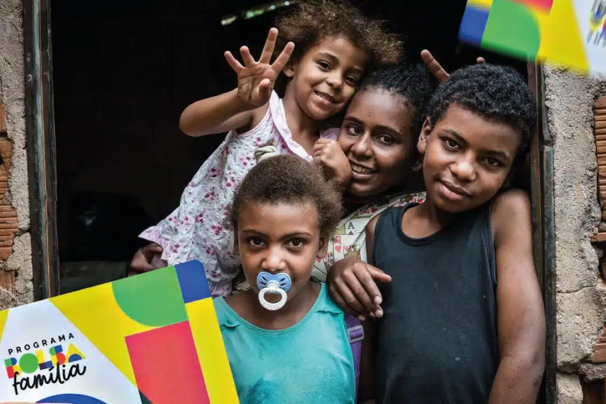 Bolsa Família Grupo de crianças e jovem em frente a uma casa simples segurando cartaz do Programa Bolsa Família