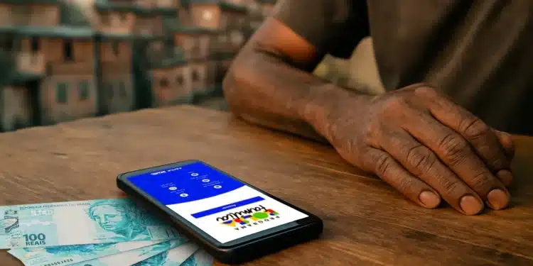 Mão de uma pessoa idosa sobre a mesa, ao lado de um smartphone com o aplicativo do Bolsa Família e cédulas de R$ 100, com uma comunidade ao fundo, simbolizando a necessidade de organização financeira.