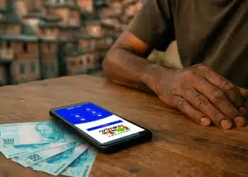 Celular com app do Bolsa Família aberto ao lado de notas de 100 reais em mesa de madeira.