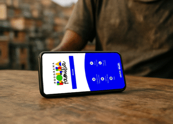 Smartphone exibindo a tela inicial do aplicativo do Bolsa Família, com opções de acesso e informações sobre o programa, em uma superfície de madeira.