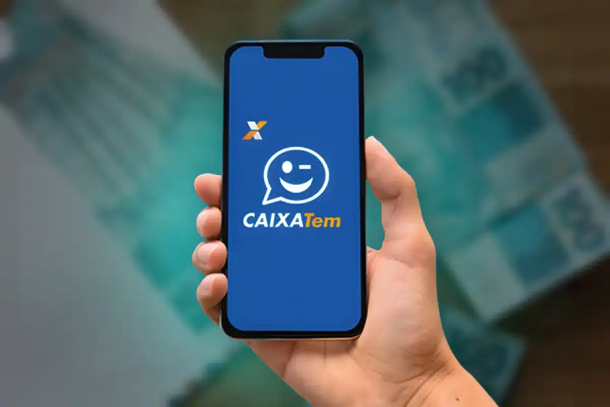 Aplicativo Caixa Tem Mão segurando um smartphone com o aplicativo Caixa Tem aberto, sobre notas de R$ 100, indicando a consulta de saldo e o acesso ao benefício.