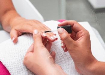 Mãos de uma pessoa sendo cuidadas por uma manicure que aplica esmalte em gel nas unhas.