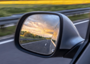 Reflexo de uma estrada e um pôr do sol com um cata-vento no retrovisor de um carro em movimento.