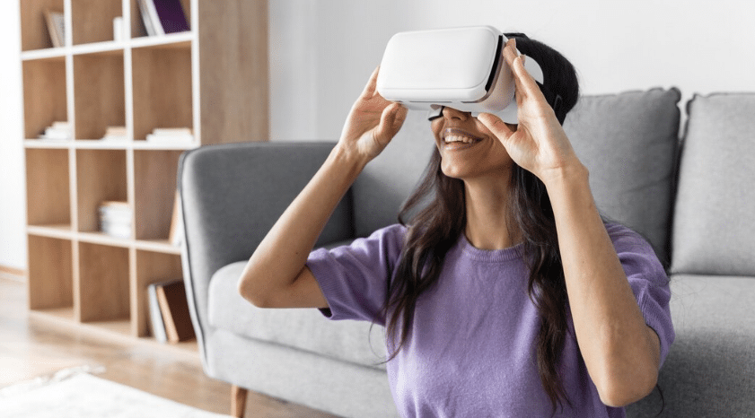 Mulher usando óculos de realidade virtual, sorrindo, em um ambiente de sala de estar. A imagem representa o uso de tecnologia avançada para treinamento e novas profissões.