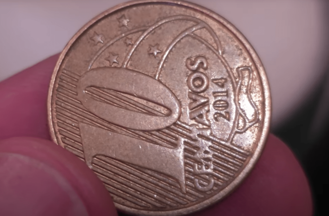 Uma moeda brasileira de 10 centavos do ano 2014, sendo segurada entre os dedos, com o reverso que exibe a inscrição "10 CENTAVOS 2014" em posição horizontal.