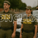 Dois policiais militares de uniforme completo em uma rua com viatura ao fundo