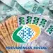 Notas de dinheiro em real com o logo da Previdência Social sobre fundo de notas de 100 e 200 reais, relacionado ao reembolso de descontos indevidos do INSS.