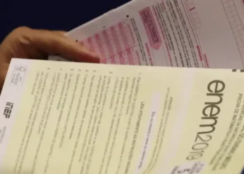 Imagem de um participante do ENEM segurando a prova, com destaque para o exame impresso e o cartão de respostas.