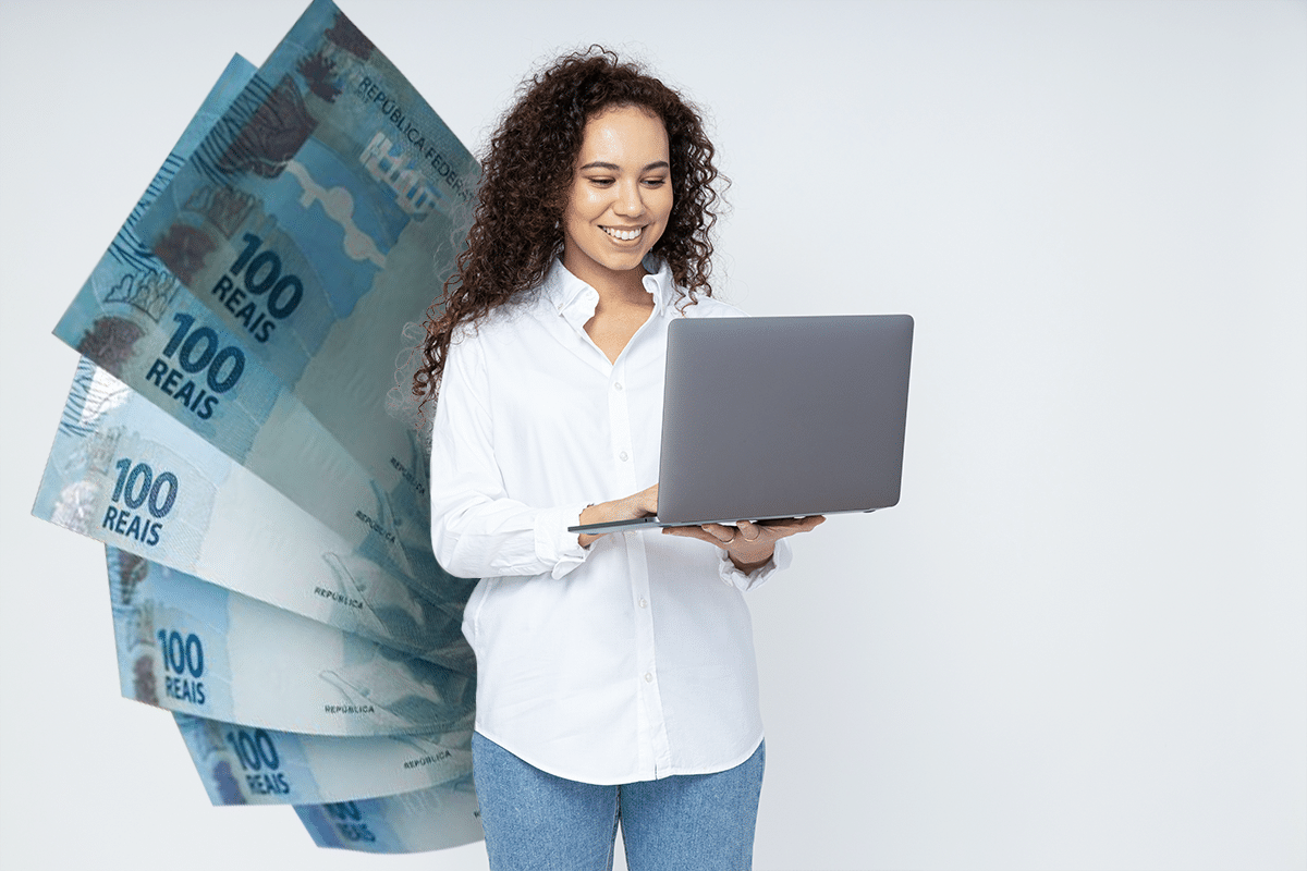 Mulher sorrindo usando laptop com cédulas de 100 reais ao fundo em parede branca