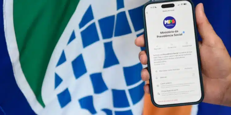 Imagem mostrando o celular com a tela do canal oficial do Ministério da Previdência Social no WhatsApp, enquanto ao fundo se vê a bandeira brasileira.