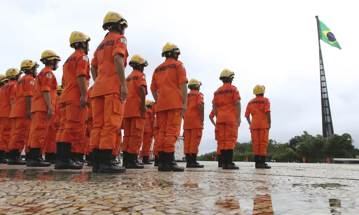 Concursos Bombeiros Grupo de bombeiros com uniforme laranja e capacete amarelo em formação, com bandeira do Brasil ao fundo