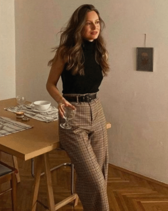 Mulher com calça xadrez marrom e blusa preta sem manga segurando taça em sala de jantar