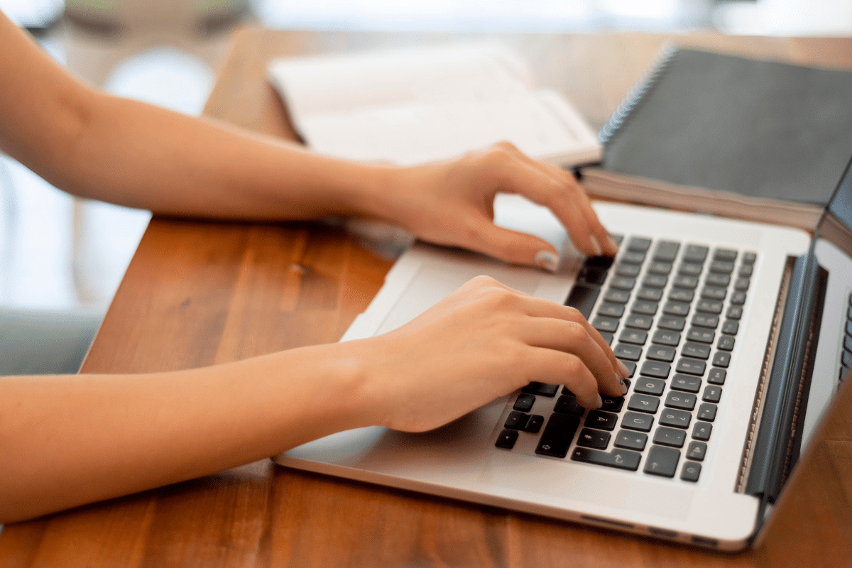 Mãos femininas digitando em teclado de notebook sobre mesa de madeira clara
