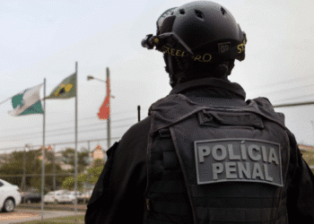 Policial penal de costas com colete tático e capacete preto em área externa com bandeiras ao fundo