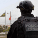 Policial penal de costas com colete tático e capacete preto em área externa com bandeiras ao fundo
