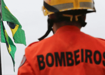 Bombeiro visto de costas usando uniforme laranja e capacete, com duas bandeiras do Brasil ao fundo