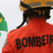 Bombeiro visto de costas usando uniforme laranja e capacete, com duas bandeiras do Brasil ao fundo