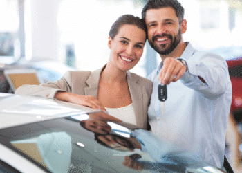 Casal sorridente segura chave ao lado de carro novo em concessionária iluminada