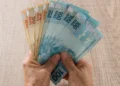 Mãos segurando cédulas de real em referência ao pagamento do BPC de setembro, conforme o final do cartão do benefício.