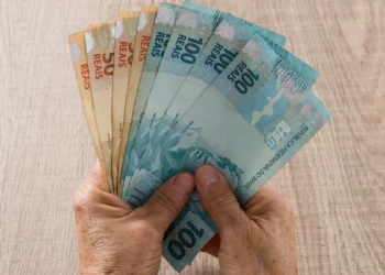 Mãos segurando cédulas de real em referência ao pagamento do BPC de setembro, conforme o final do cartão do benefício.
