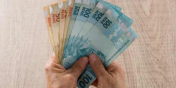 Mãos segurando cédulas de real em referência ao pagamento do BPC de setembro, conforme o final do cartão do benefício.