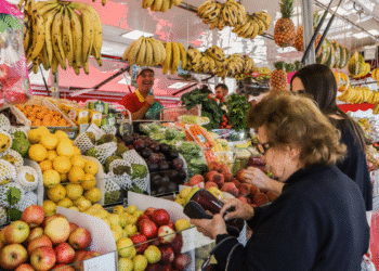 Vendedora sorridente com frutas variadas em mercado aberto e clientes escolhendo produtos