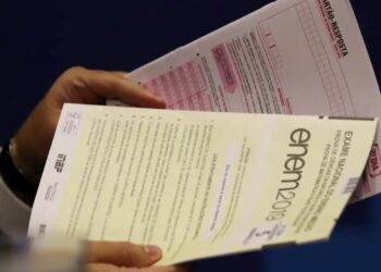 Um estudante segura duas folhas de prova do Enem 2019: um caderno de questões do segundo dia (matemática e ciências da natureza) e um cartão-resposta. O foco está nos documentos, que representam o momento da prova.