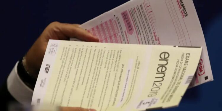 Um estudante segura duas folhas de prova do Enem 2019: um caderno de questões do segundo dia (matemática e ciências da natureza) e um cartão-resposta. O foco está nos documentos, que representam o momento da prova.