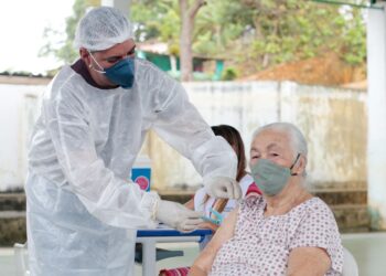 Profissional de saúde com máscara e roupa de proteção aplicando vacina em braço de idosa com máscara.