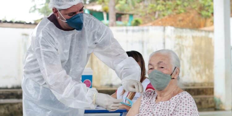 Profissional de saúde com máscara e roupa de proteção aplicando vacina em braço de idosa com máscara.