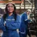 Mulher sorrindo em uniforme de trabalho industrial, com capacete e óculos de proteção, em um ambiente de fábrica, enquanto um homem trabalha ao fundo, ambos com roupas de segurança.