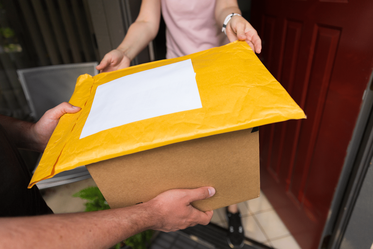 Mãos trocando um pacote de papelão e um envelope amarelo na porta de uma casa