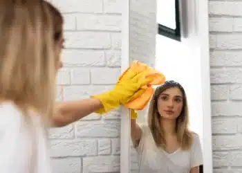 Mulher limpando espelho com pano, usando luvas amarelas, para deixar sem manchas.