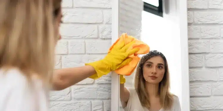 Mulher limpando espelho com pano, usando luvas amarelas, para deixar sem manchas.