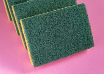 Esponjas de cozinha novas em close, com a parte amarela e a fibra verde visíveis, sobre um fundo rosa.